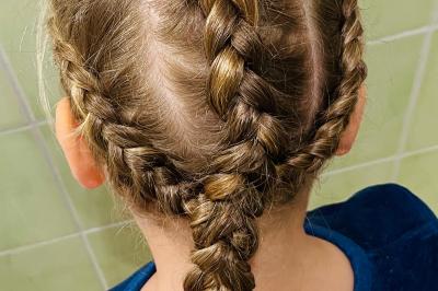 Femme de dos avec des cheveux blonds coiffés en tresse et chignon, portant un pull beige près d'une fenêtre.