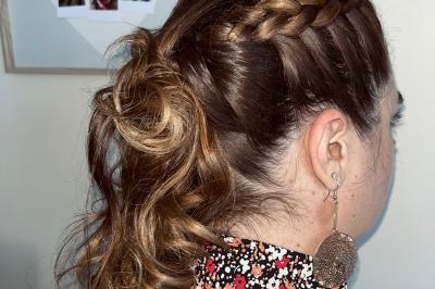 Femme de dos avec des cheveux blonds coiffés en tresse et chignon, portant un pull beige près d'une fenêtre.