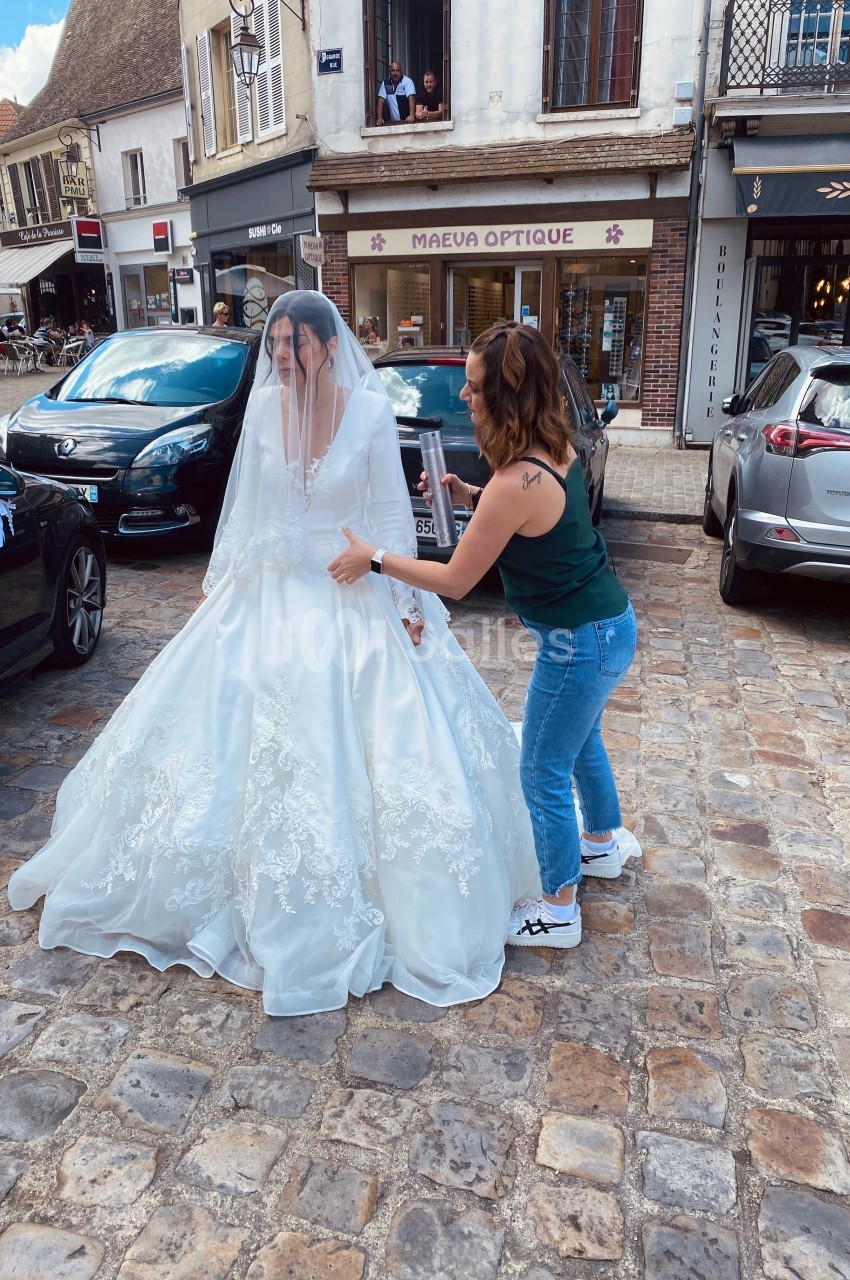 Une femme ajuste la robe de mariée d'une autre femme portant un voile, sur une place pavée bordée de boutiques.