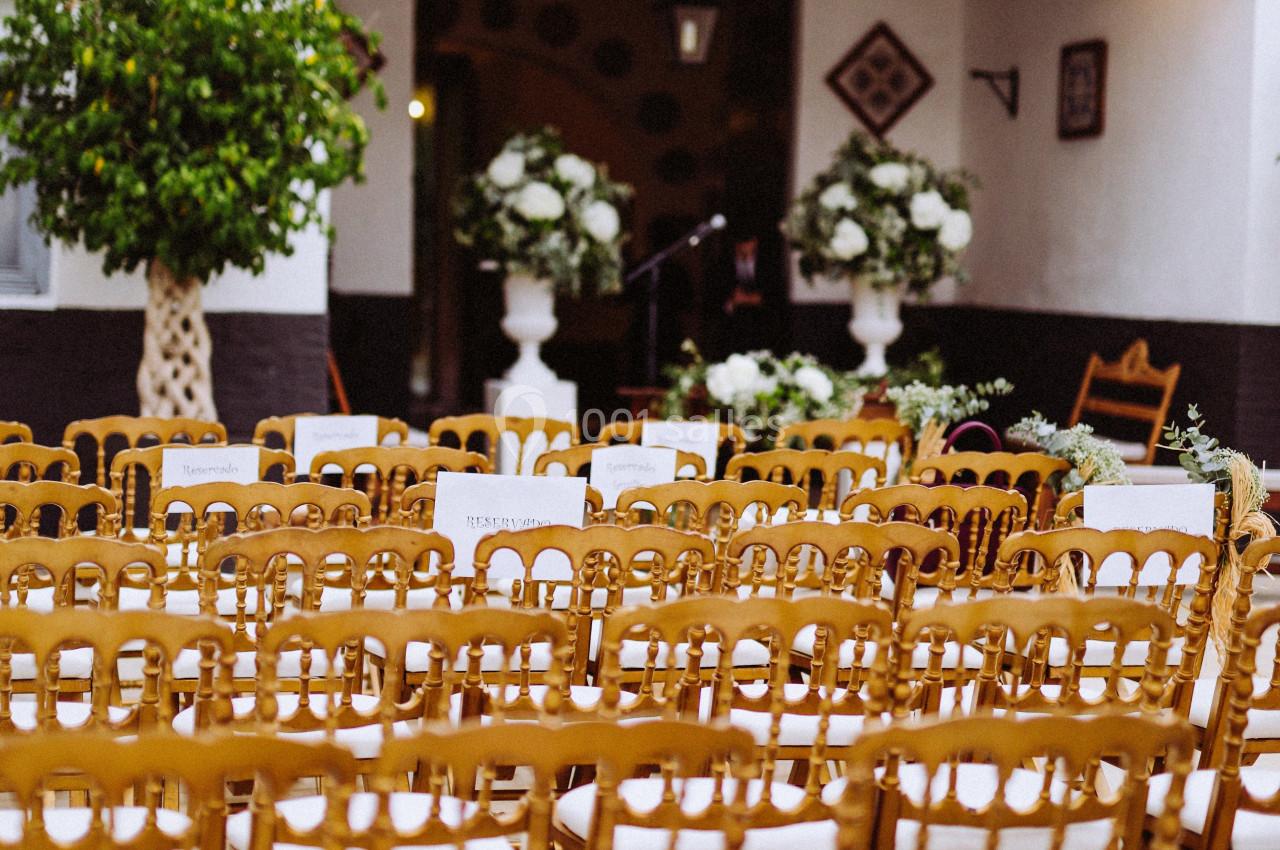 Chaises en bois alignées devant une arche décorée de fleurs blanches dans une cour extérieure.