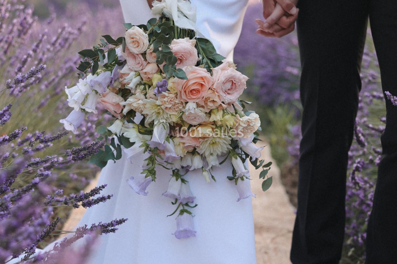 Un couple en tenue de mariage se tient la main dans un champ de lavande, avec un bouquet de fleurs pastel.