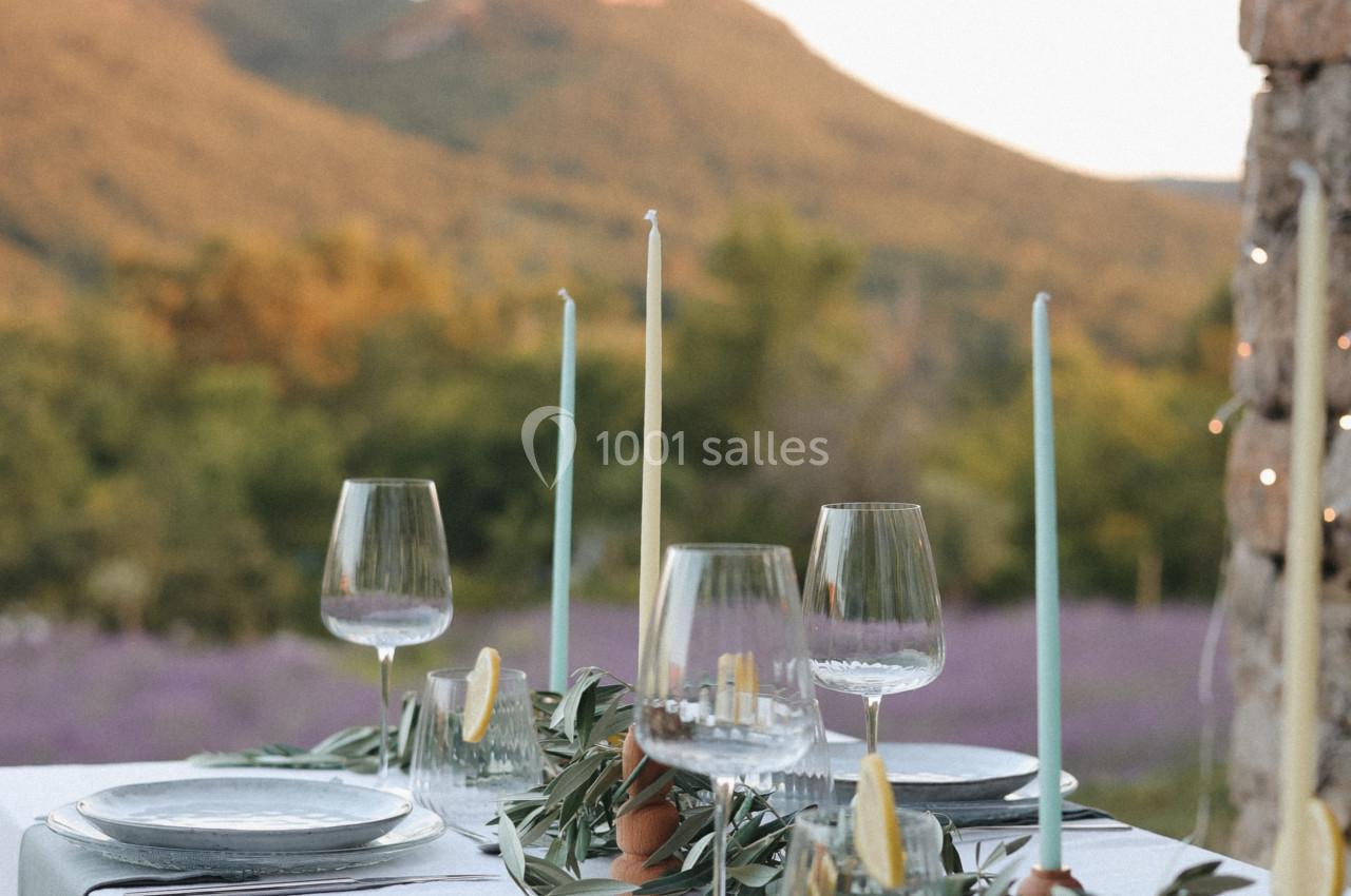 Table dressée en extérieur avec nappes blanches, bougies, verres à pied et décorations végétales devant un paysage…