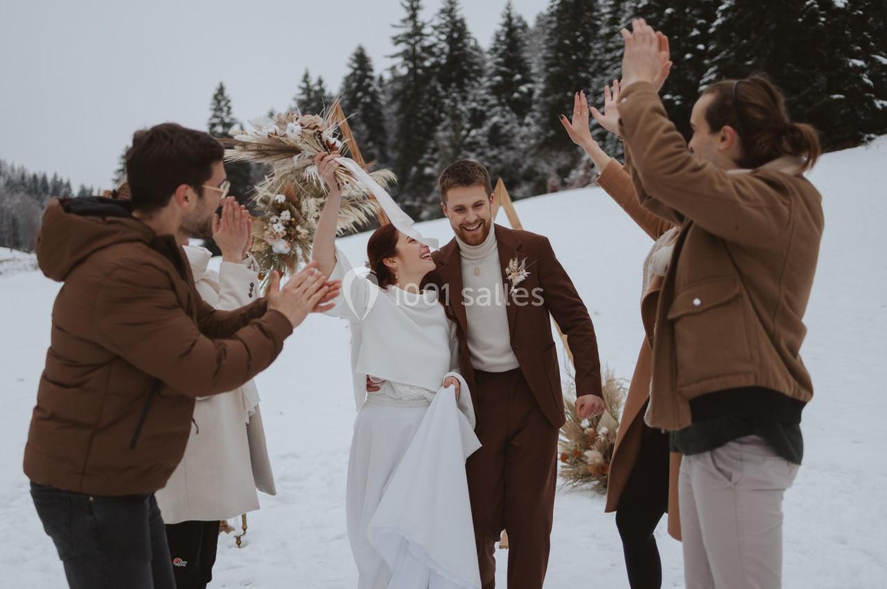 Un couple célèbre son mariage en extérieur sous la neige, entouré de proches applaudissant joyeusement.