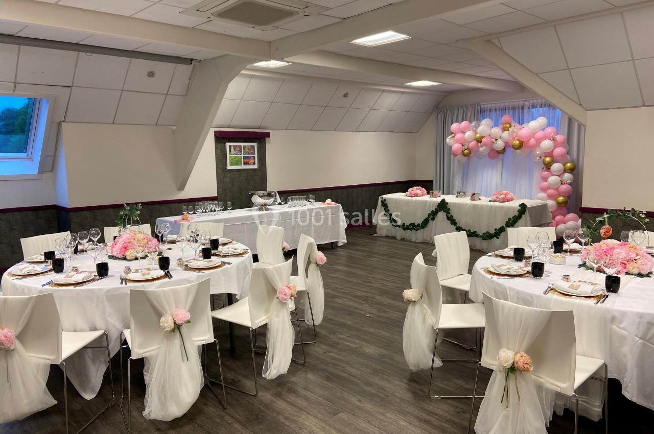 Salle décorée pour un événement, avec tables rondes ornées de fleurs roses et arche de ballons en arrière-plan.