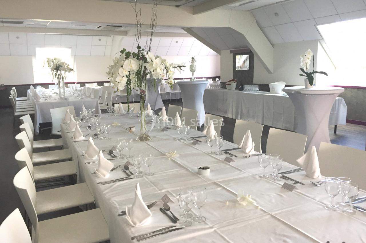 Salle de réception décorée avec des tables dressées en blanc, ornées de fleurs et de serviettes pliées.