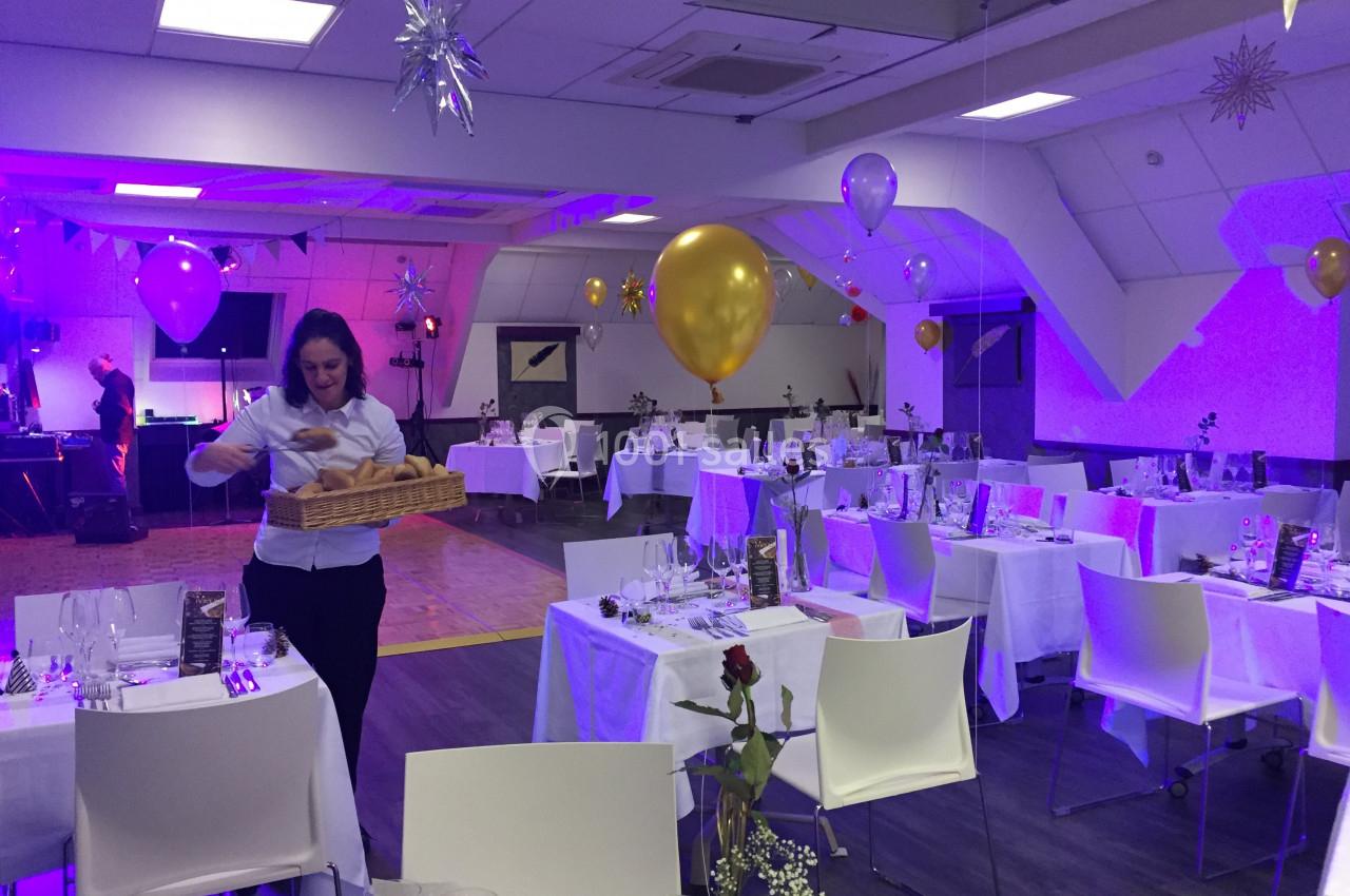 Salle de réception décorée avec ballons et tables dressées, une personne prépare des paniers de pain.