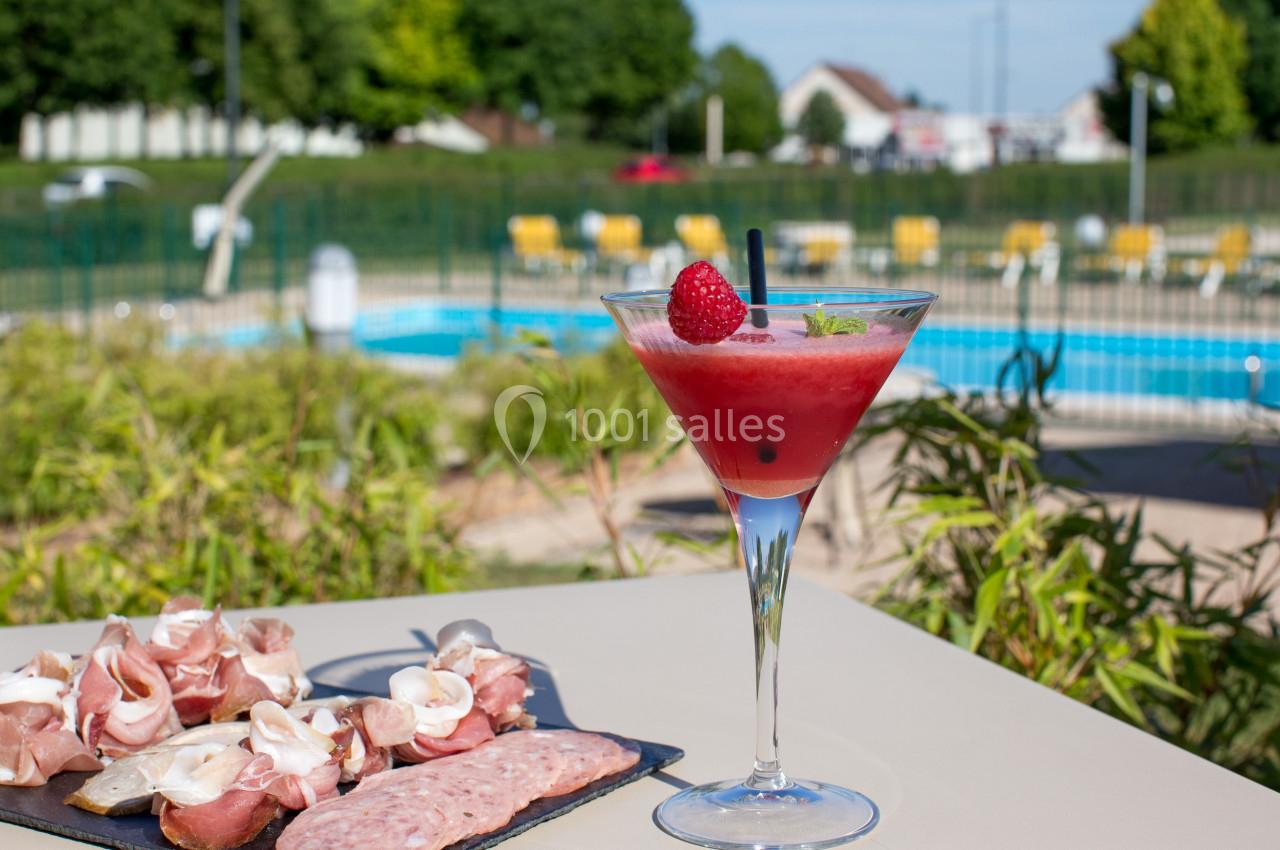 Cocktail rouge garni d'une framboise et plateau de charcuterie sur une table près d'une piscine en extérieur.
