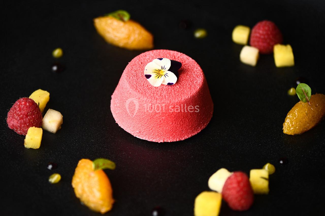 Dessert rose texturé garni d'une fleur comestible, entouré de fruits frais et de petites touches de sauce sur une assiette…