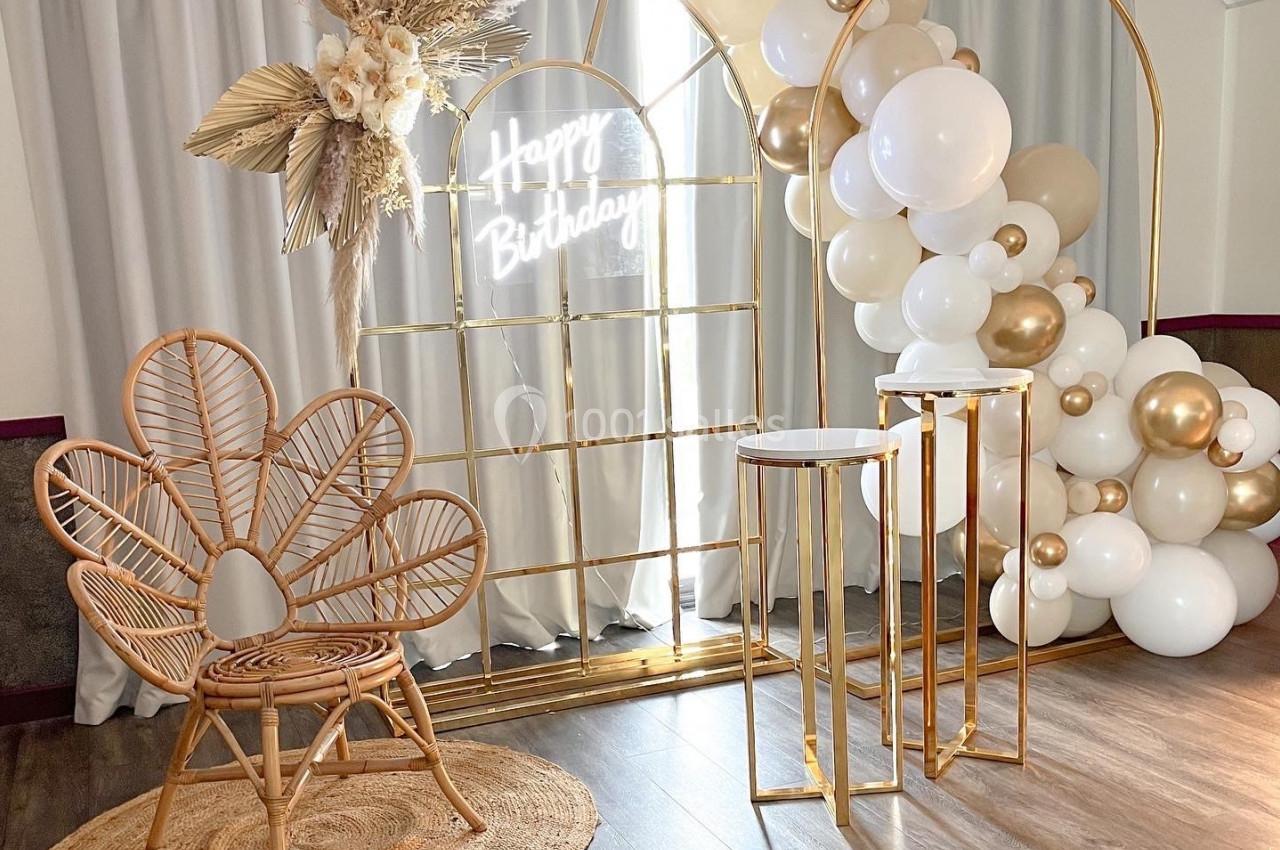 Décor d'anniversaire avec arche dorée, ballons blancs et dorés, chaise en rotin et fleurs séchées.
