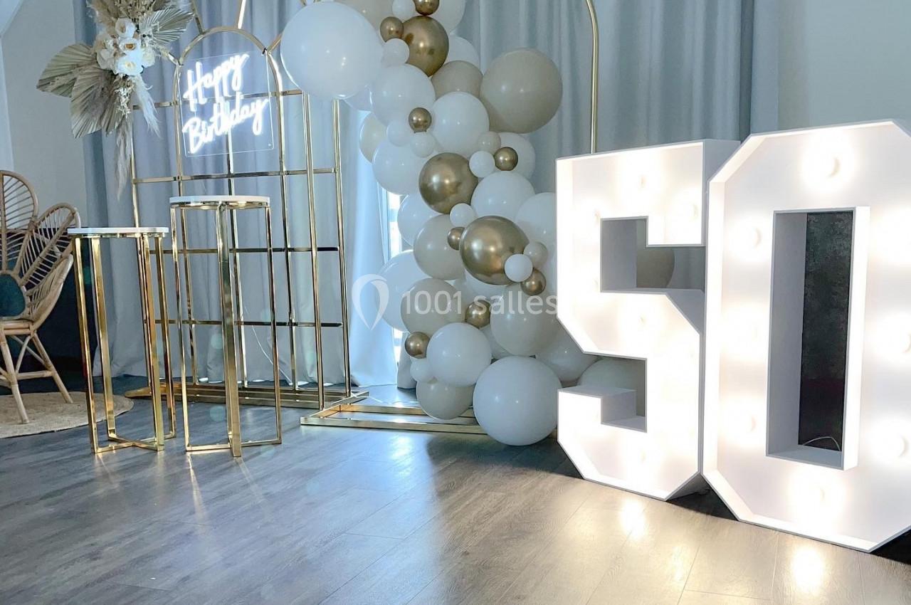 Décoration d'anniversaire avec ballons blancs et dorés, arche métallique, et chiffres lumineux ’50’ sur un sol en bois.