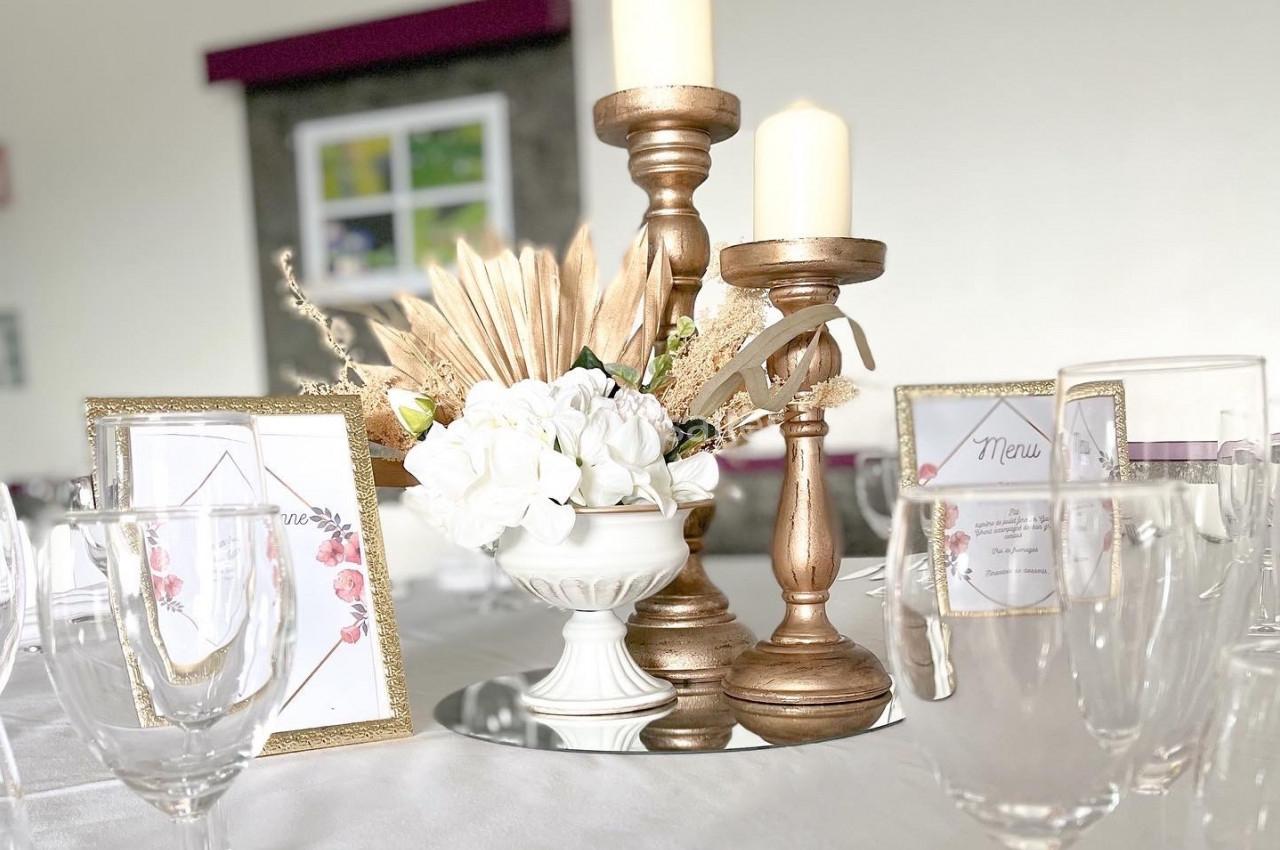 Centre de table avec chandeliers dorés, bougies, fleurs blanches et cadres décoratifs sur une nappe blanche.