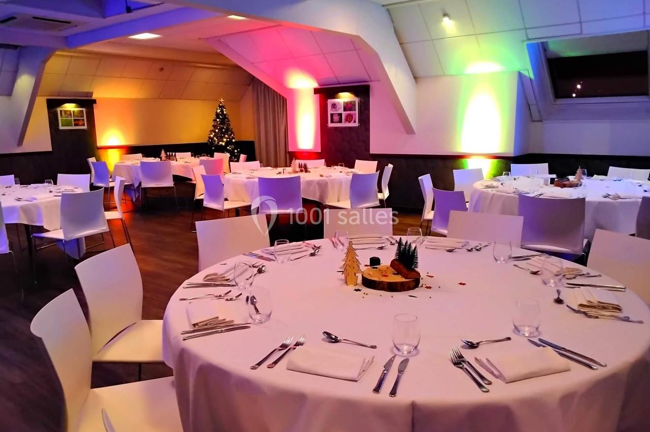 Salle de réception décorée avec des tables rondes dressées, éclairage coloré et sapin de Noël en arrière-plan.