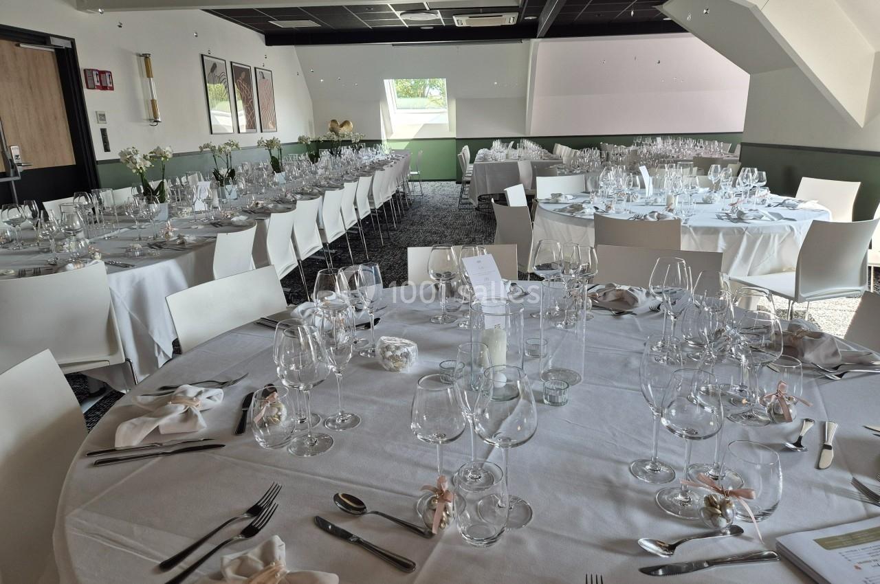 Salle de réception lumineuse avec des tables rondes dressées pour un repas, ornées de verres et décorations élégantes.