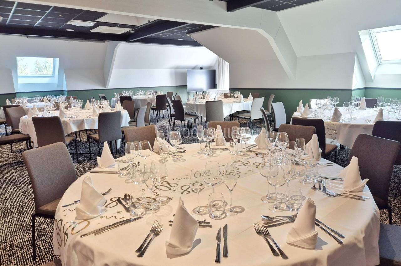 Salle de réception avec tables rondes dressées, nappes blanches, verres et couverts, éclairée par des fenêtres de toit.