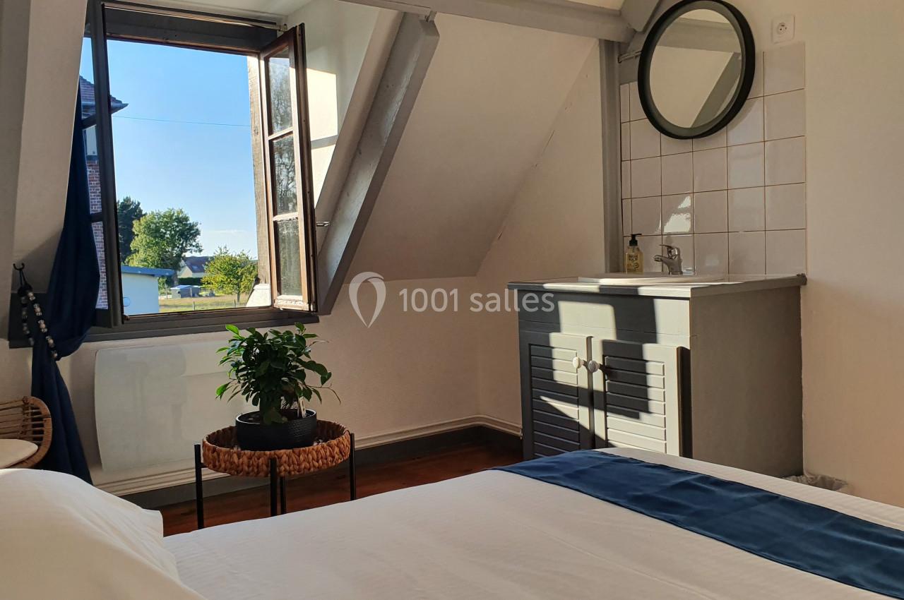 Chambre lumineuse avec un lit, une fenêtre ouverte donnant sur l'extérieur, un lavabo et une plante sur une table.