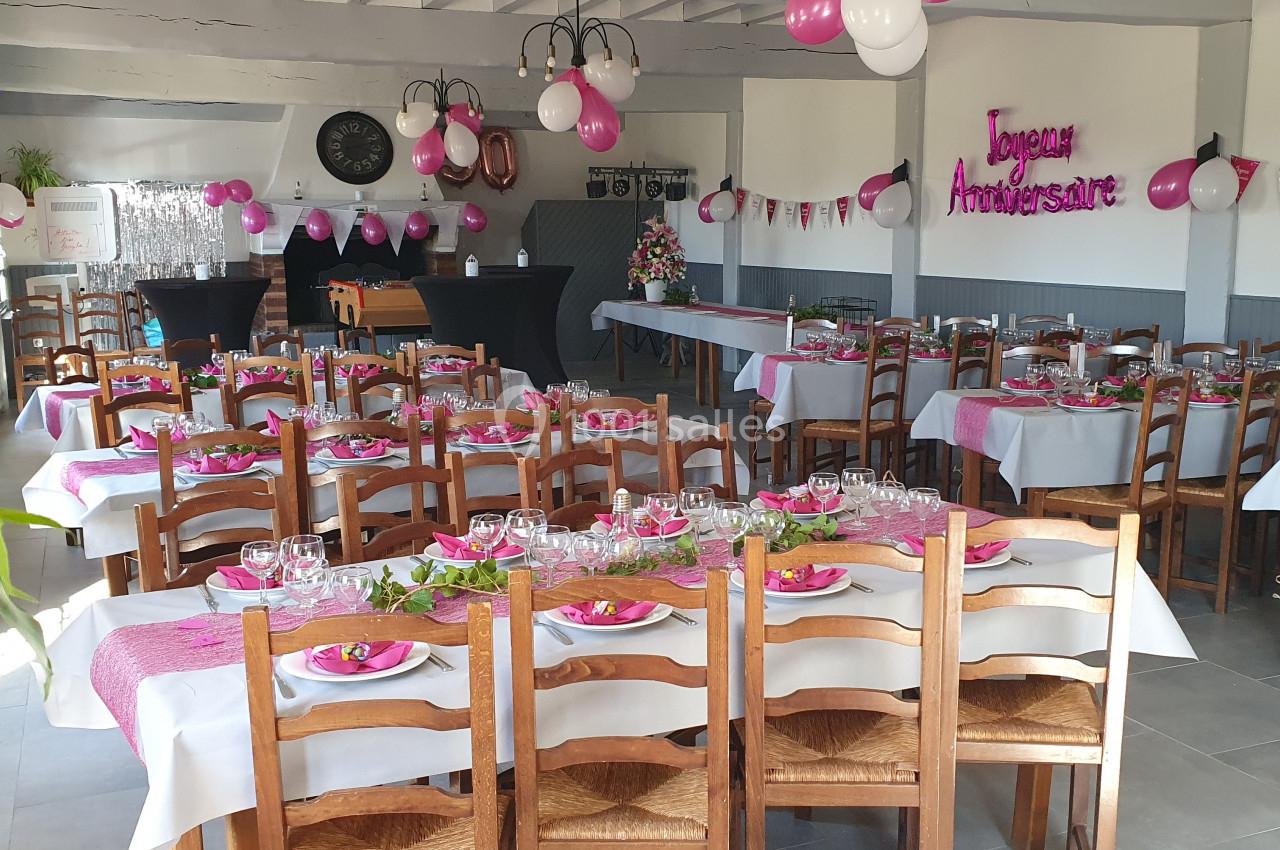 Salle décorée pour un anniversaire avec des tables dressées, des ballons roses et blancs, et une banderole ’Joyeux…