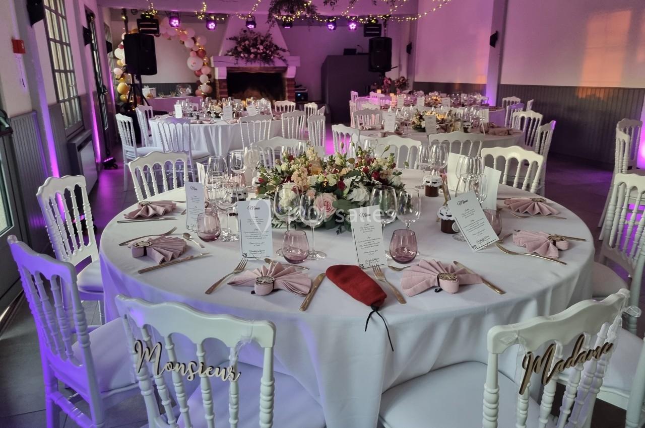 Salle de réception décorée pour un mariage, avec tables rondes dressées, fleurs, menus et éclairage tamisé.