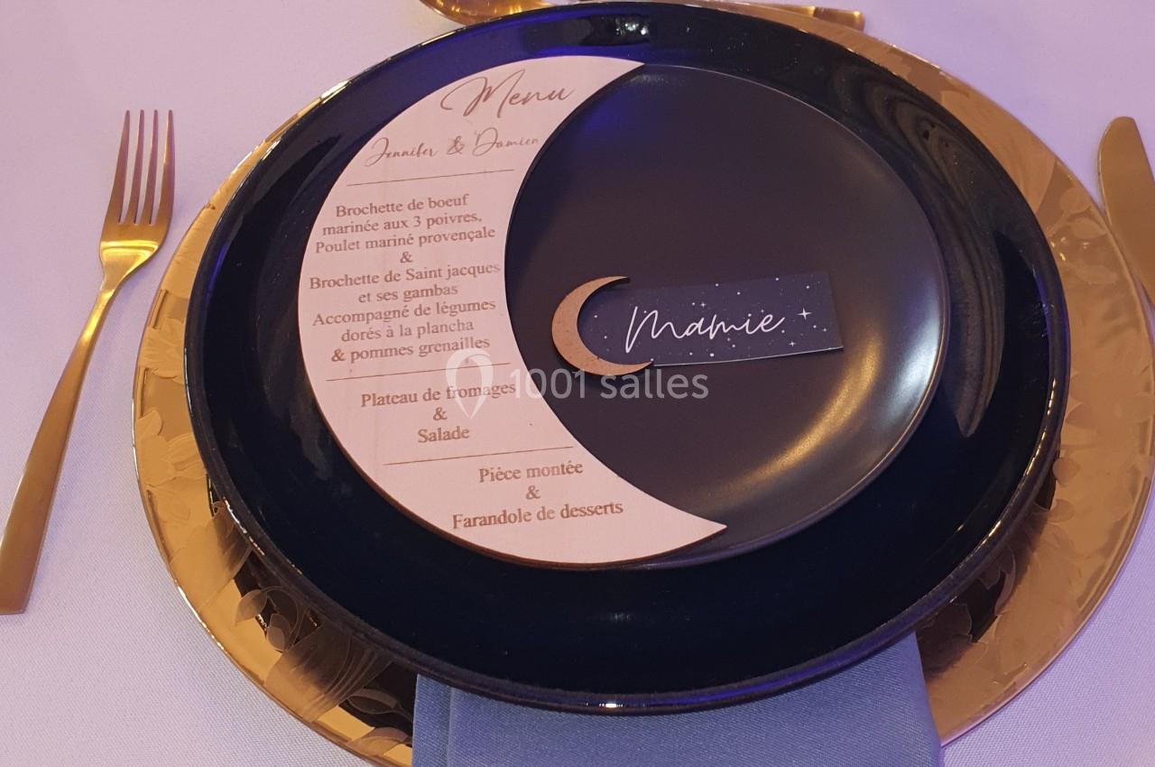 Assiette noire avec un menu imprimé et une serviette bleue, disposées sur une table dressée avec des couverts dorés.