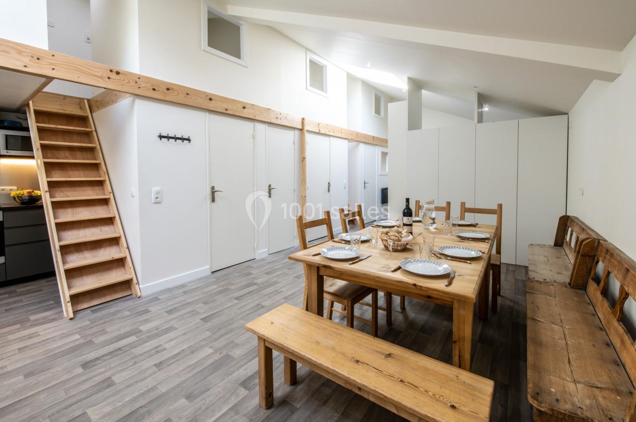 Salle à manger lumineuse avec table en bois dressée, bancs assortis, escalier en bois et cuisine visible en arrière-plan.