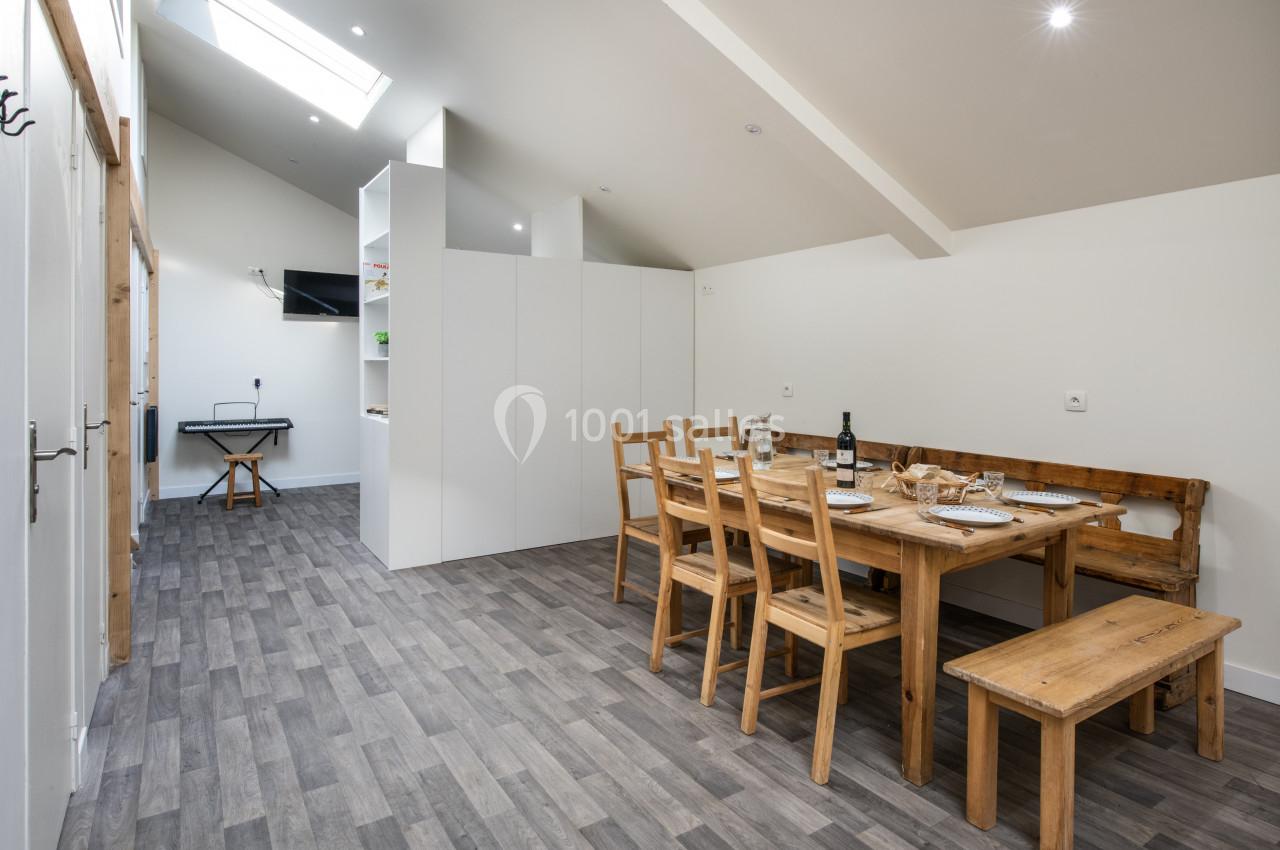 Salle à manger lumineuse avec table en bois, chaises assorties, sol en parquet gris et cuisine ouverte en arrière-plan.