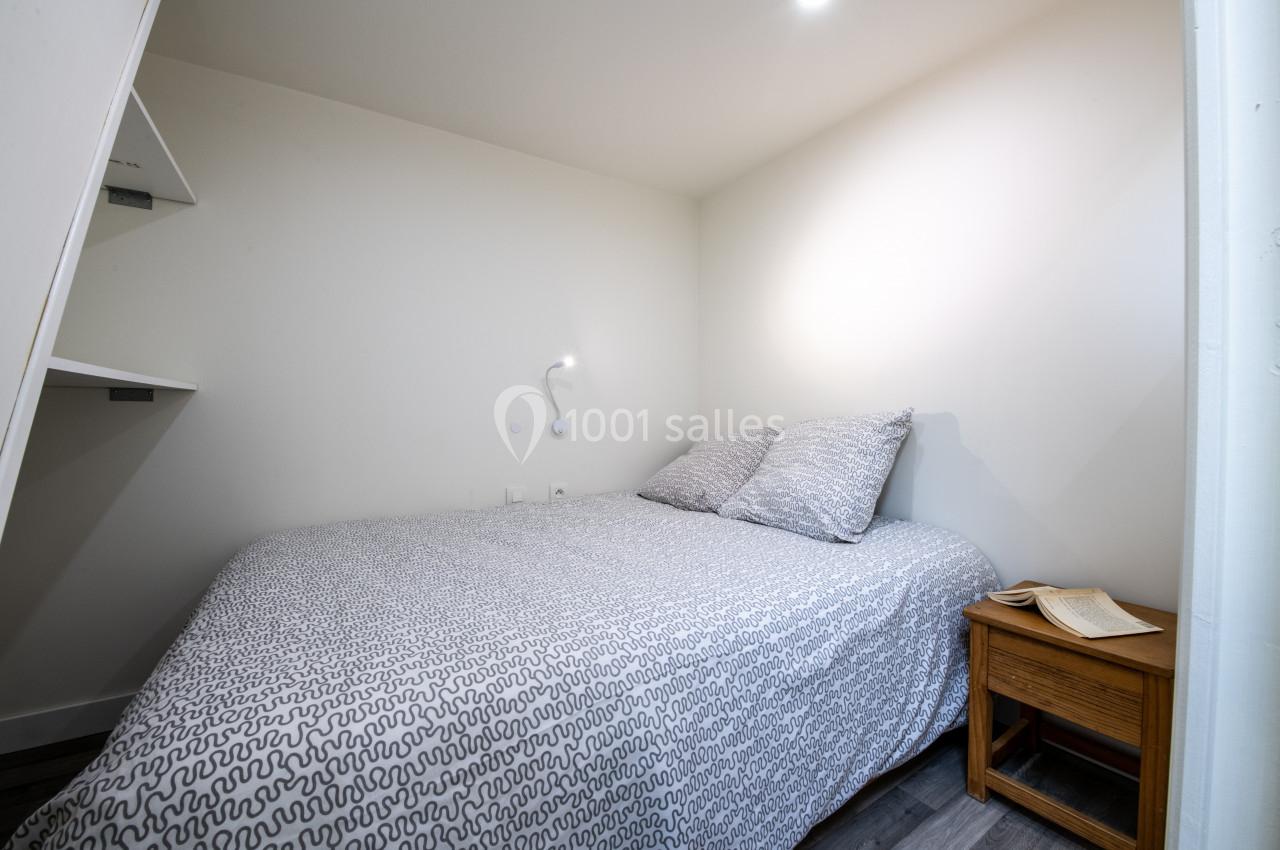 Lit double avec housse de couette grise, table de chevet en bois avec un livre ouvert, dans une petite chambre lumineuse.