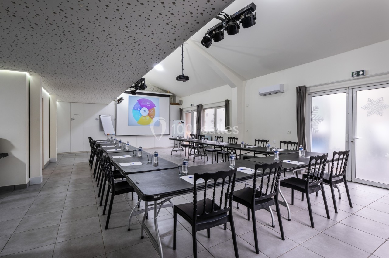 Salle de réunion lumineuse avec tables disposées en U, écran de projection allumé et chaises noires.