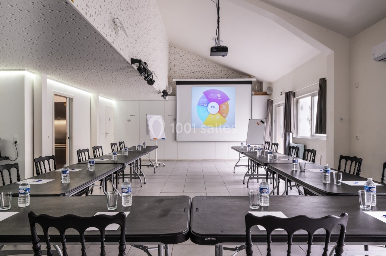 Salle de réunion lumineuse avec tables disposées en U, écran de projection, paperboards et bouteilles d'eau.