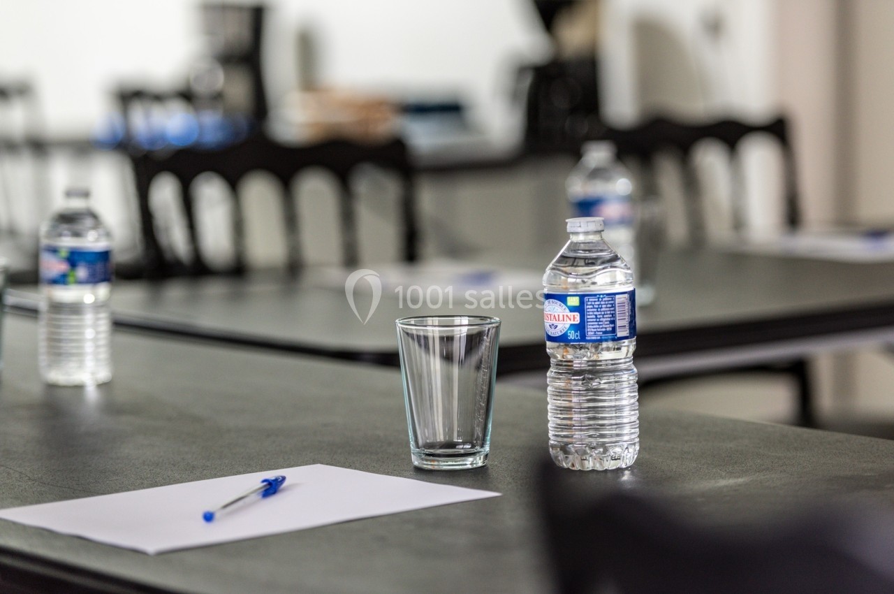 Bouteille d'eau, verre vide, stylo et feuille posés sur une table dans une salle avec des chaises en arrière-plan.
