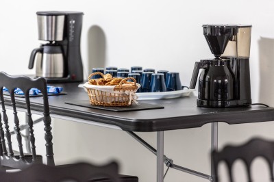 Table avec cafetières, tasses bleues empilées et panier de viennoiseries, dans un espace de pause.