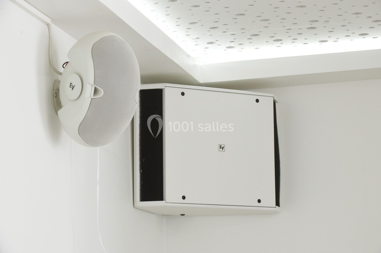 Haut-parleur mural blanc fixé dans un angle, avec un plafond perforé visible en arrière-plan.