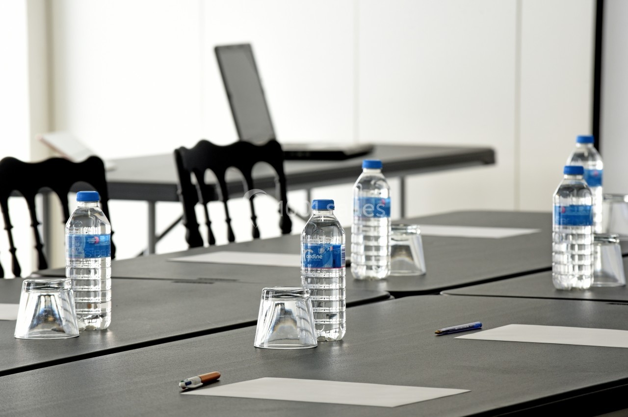 Salle de réunion avec tables noires, bouteilles d'eau, verres, stylos et feuilles, ordinateur en arrière-plan.