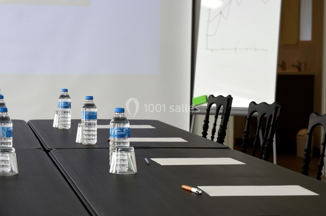 Salle de réunion avec table noire, bouteilles d'eau, feuilles blanches, stylos et tableau blanc en arrière-plan.