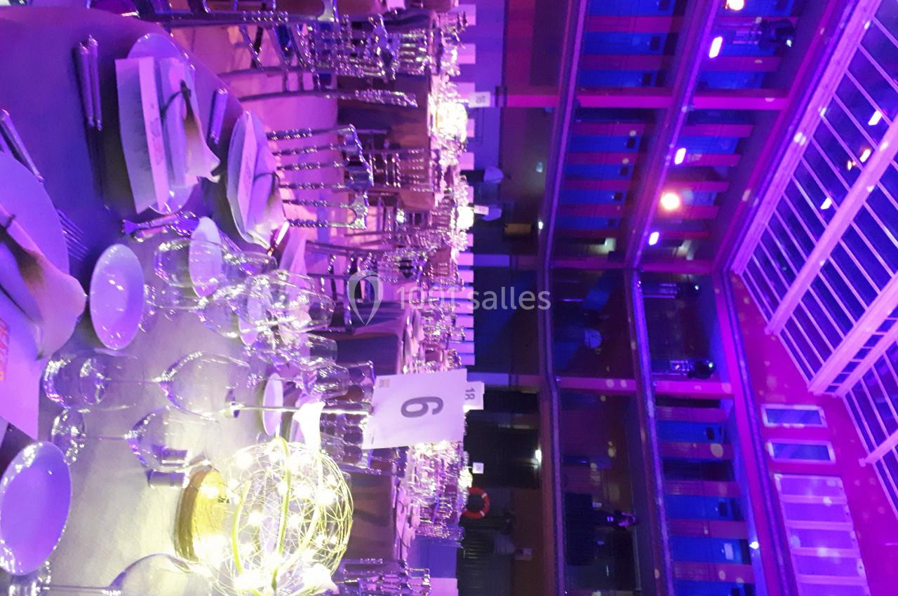 Salle de réception illuminée en violet avec tables dressées, chaises en métal et centre de table lumineux.