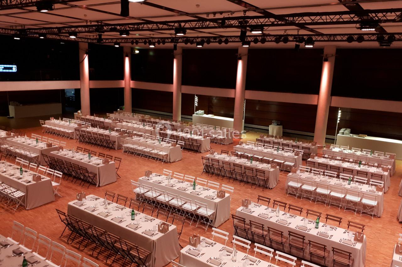 Salle de réception avec de longues tables dressées pour un événement, éclairage chaleureux et disposition ordonnée.
