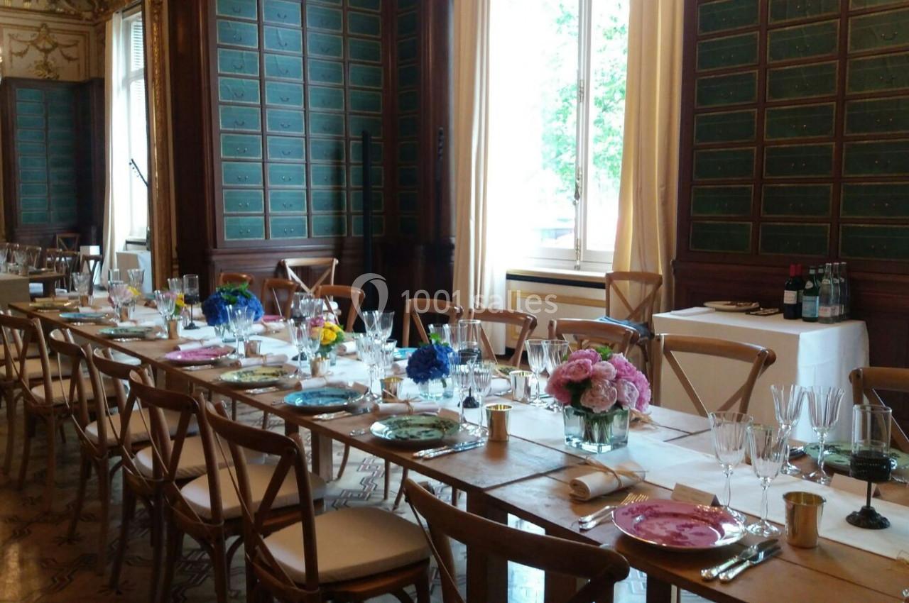 Table élégamment dressée dans une salle ornée de boiseries et de grandes fenêtres, avec des fleurs et de la vaisselle…