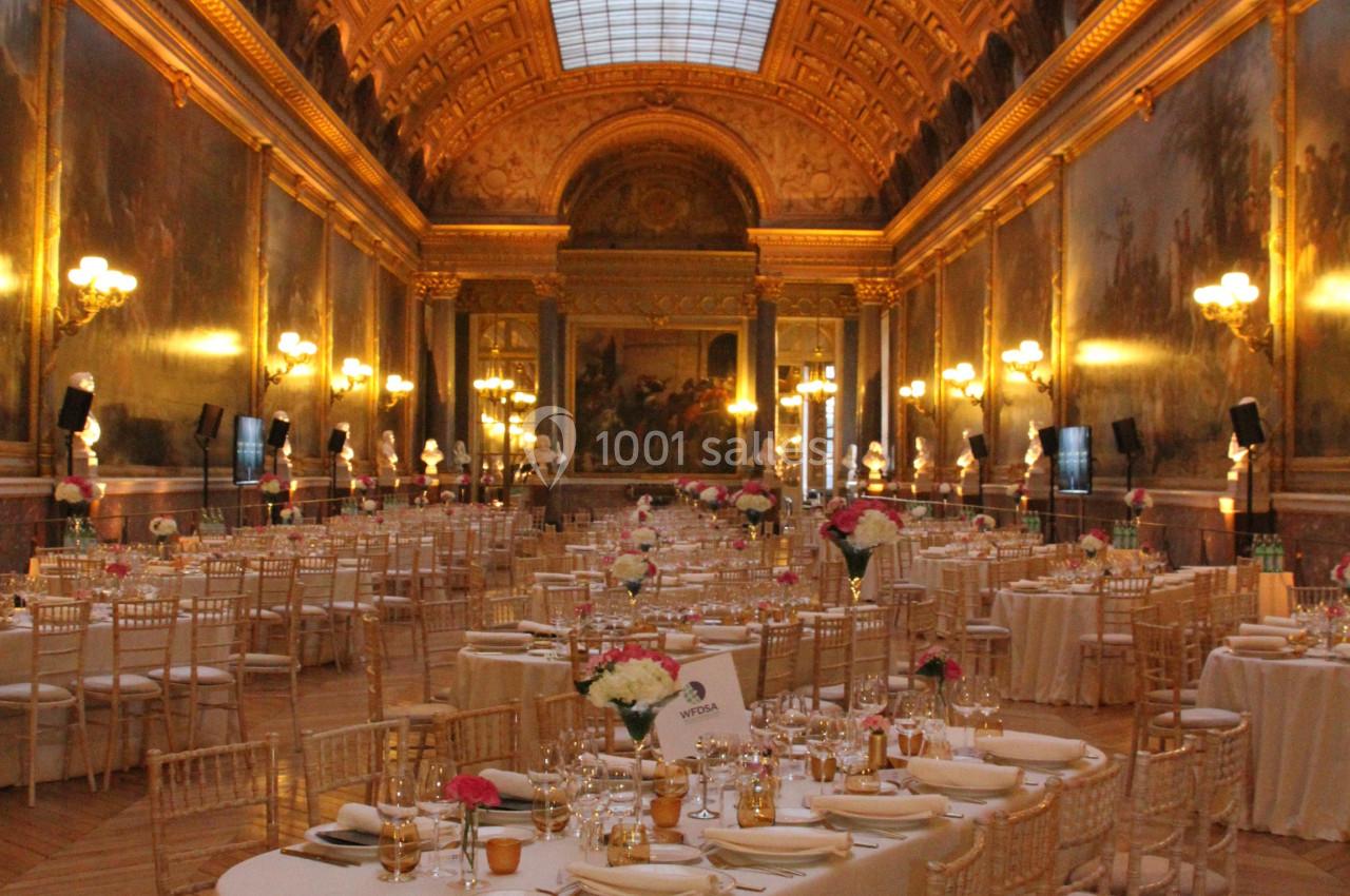 Salle de réception élégante avec tables rondes dressées, éclairée par des lustres et un plafond en verrière.