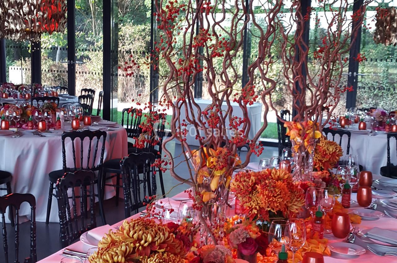 Table décorée pour un événement avec des fleurs orange, branches torsadées et vaisselle élégante dans une salle vitrée.