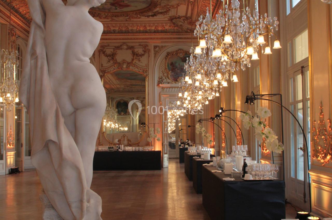 Statue en marbre dans une salle ornée de lustres, fresques au plafond et tables de réception alignées.