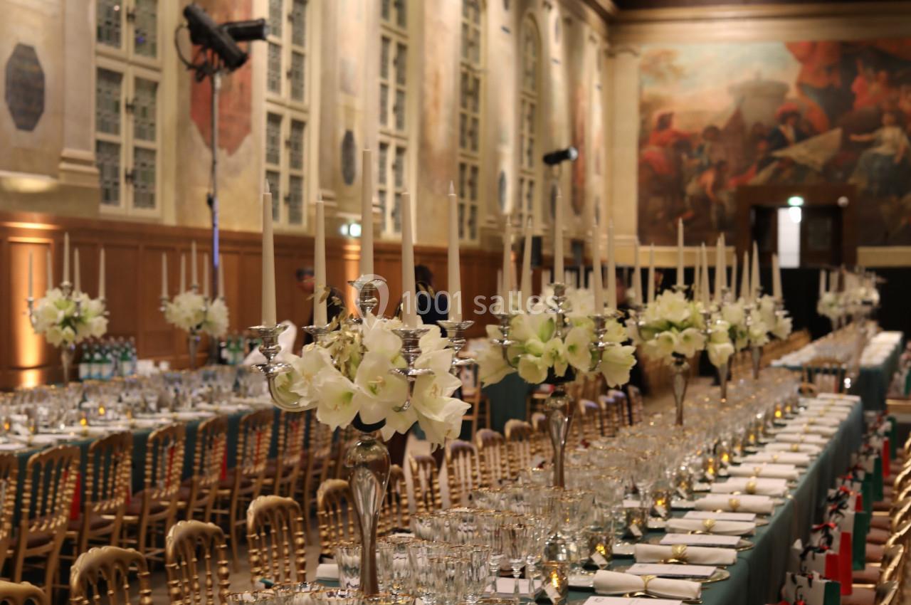 Salle de réception élégante avec longues tables dressées, chandeliers, fleurs blanches et décorations raffinées.