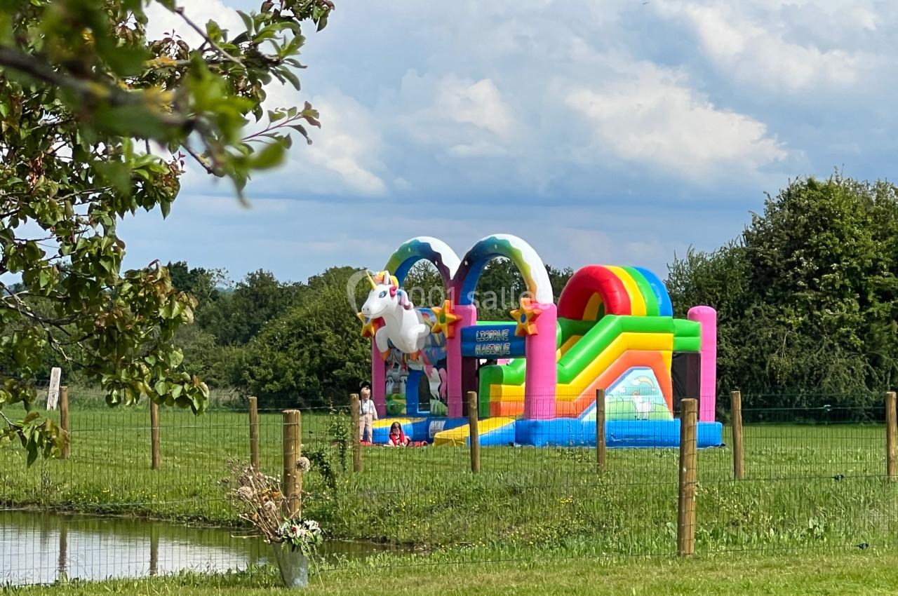 Structure gonflable colorée en forme de licorne dans un champ verdoyant, avec des enfants jouant à proximité.