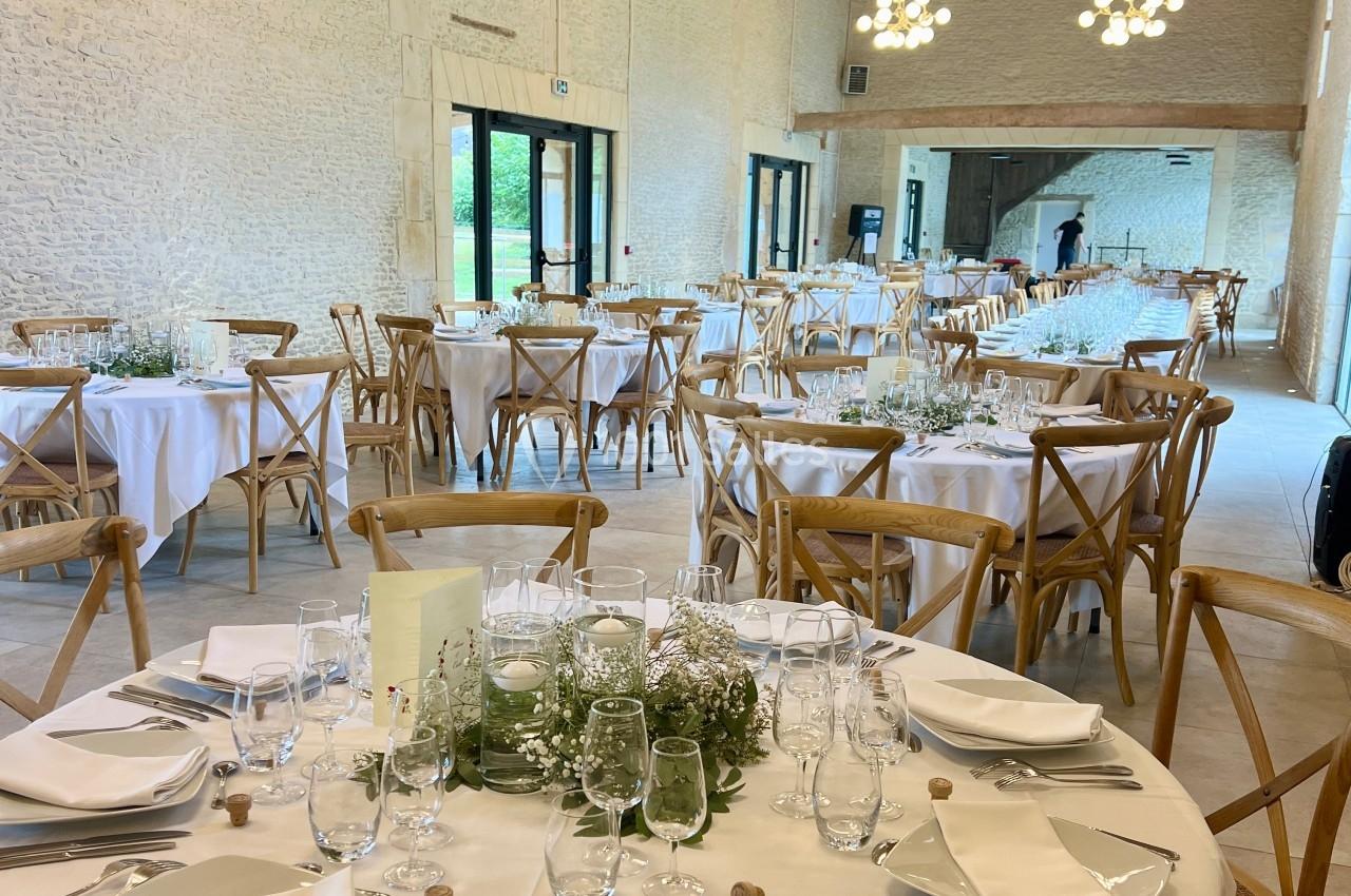 Salle de réception lumineuse avec tables rondes dressées, nappes blanches et décorations florales.