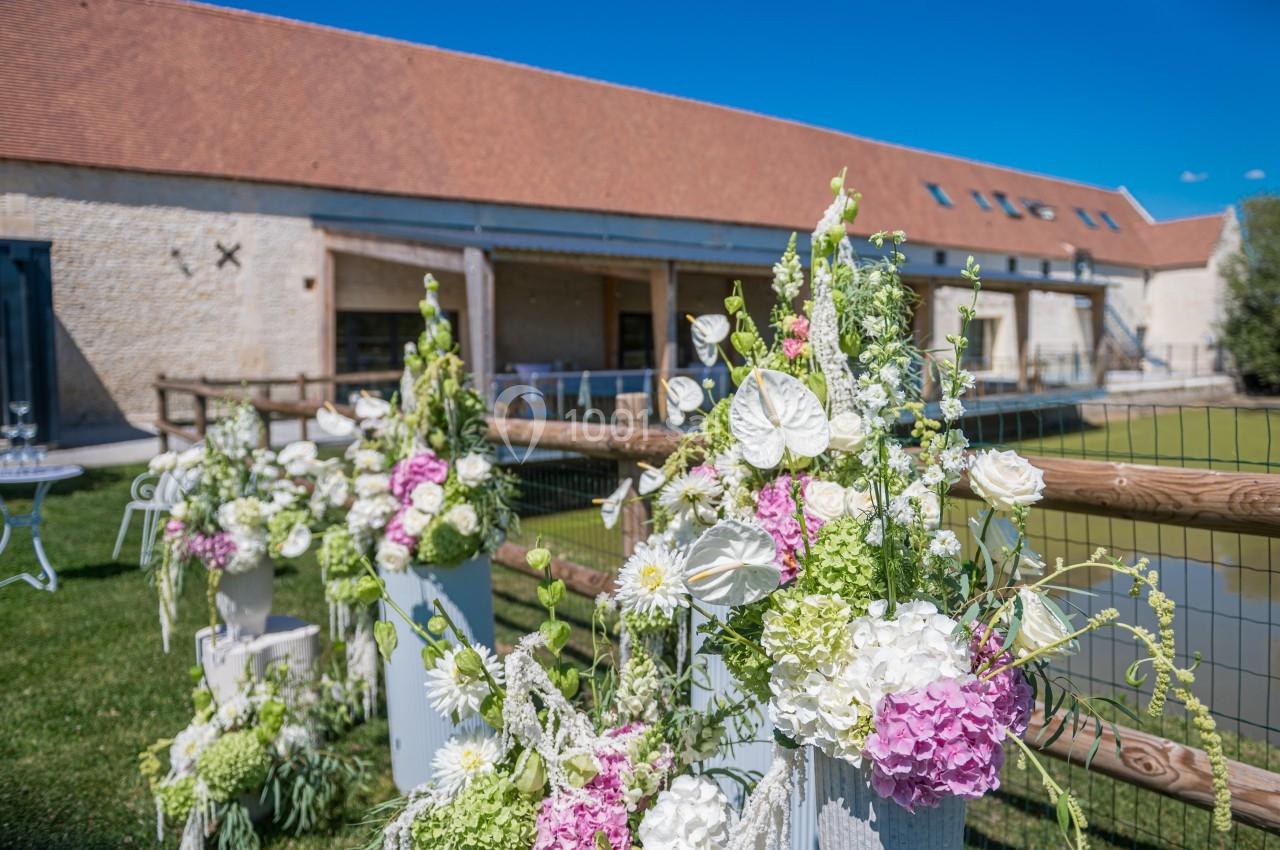 Décoration florale blanche et rose sur des supports devant un bâtiment en pierre avec toit en tuiles, par temps ensoleillé.