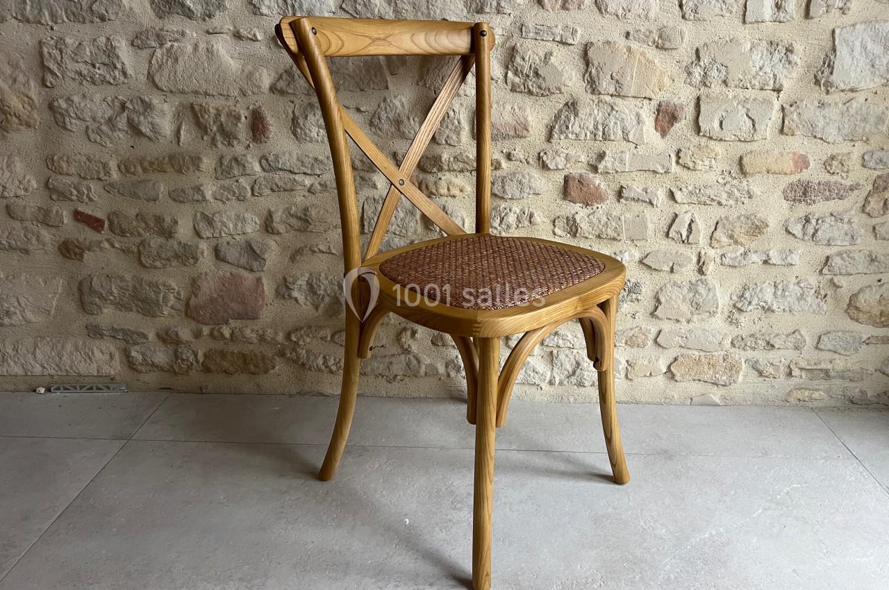 Chaise en bois clair avec dossier croisé et assise en rotin, posée sur un sol en carrelage devant un mur en pierre.