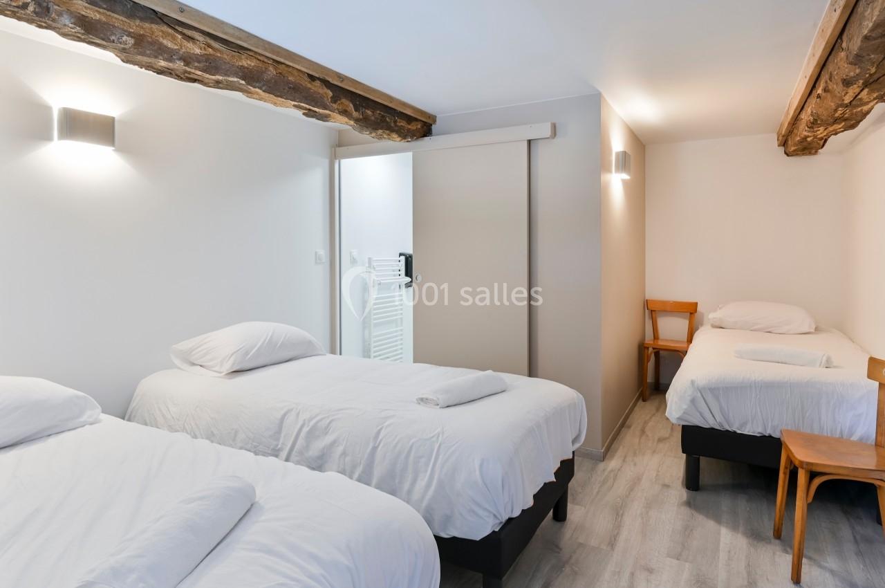 Chambre lumineuse avec trois lits simples, murs blancs, poutres apparentes et sol en parquet clair.