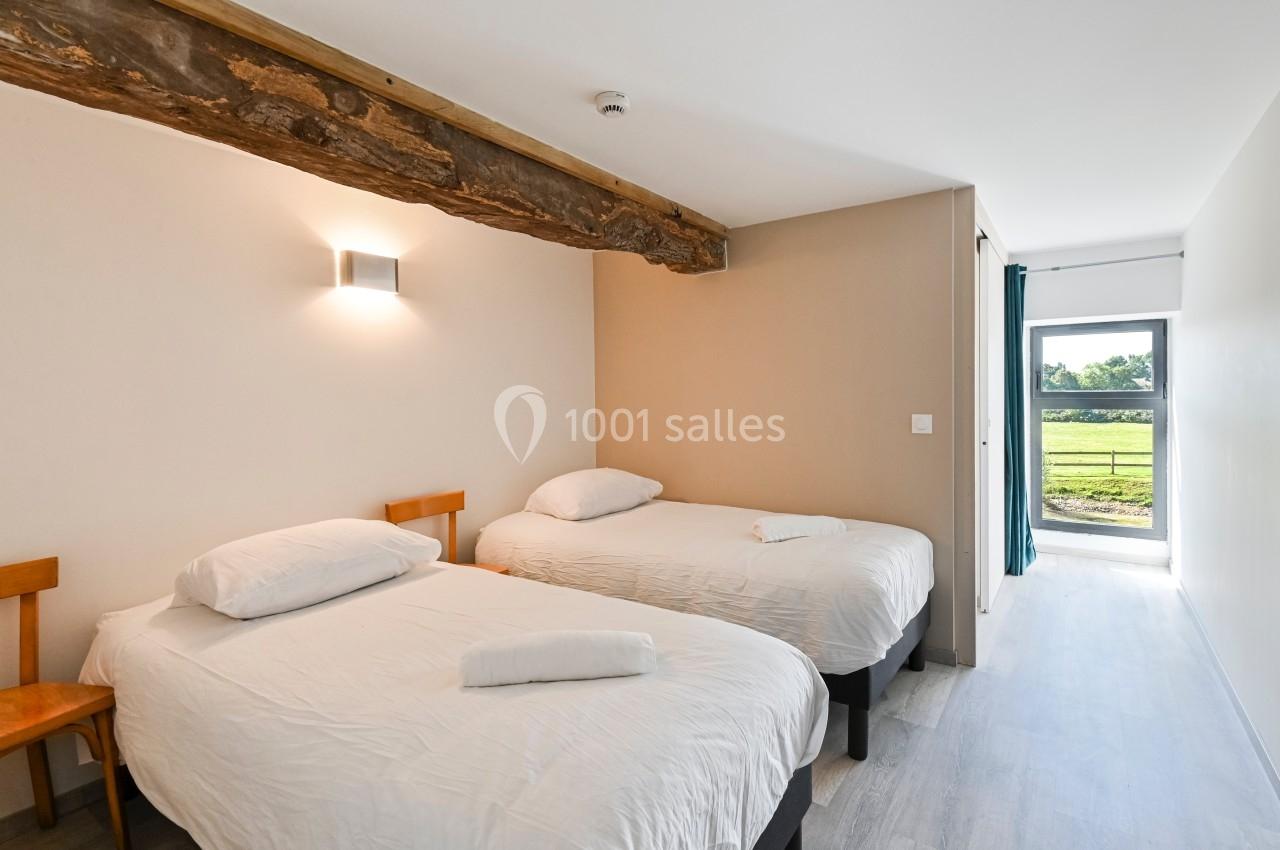 Deux lits simples dans une chambre lumineuse avec poutre apparente, sol clair et vue sur un espace vert par une porte…