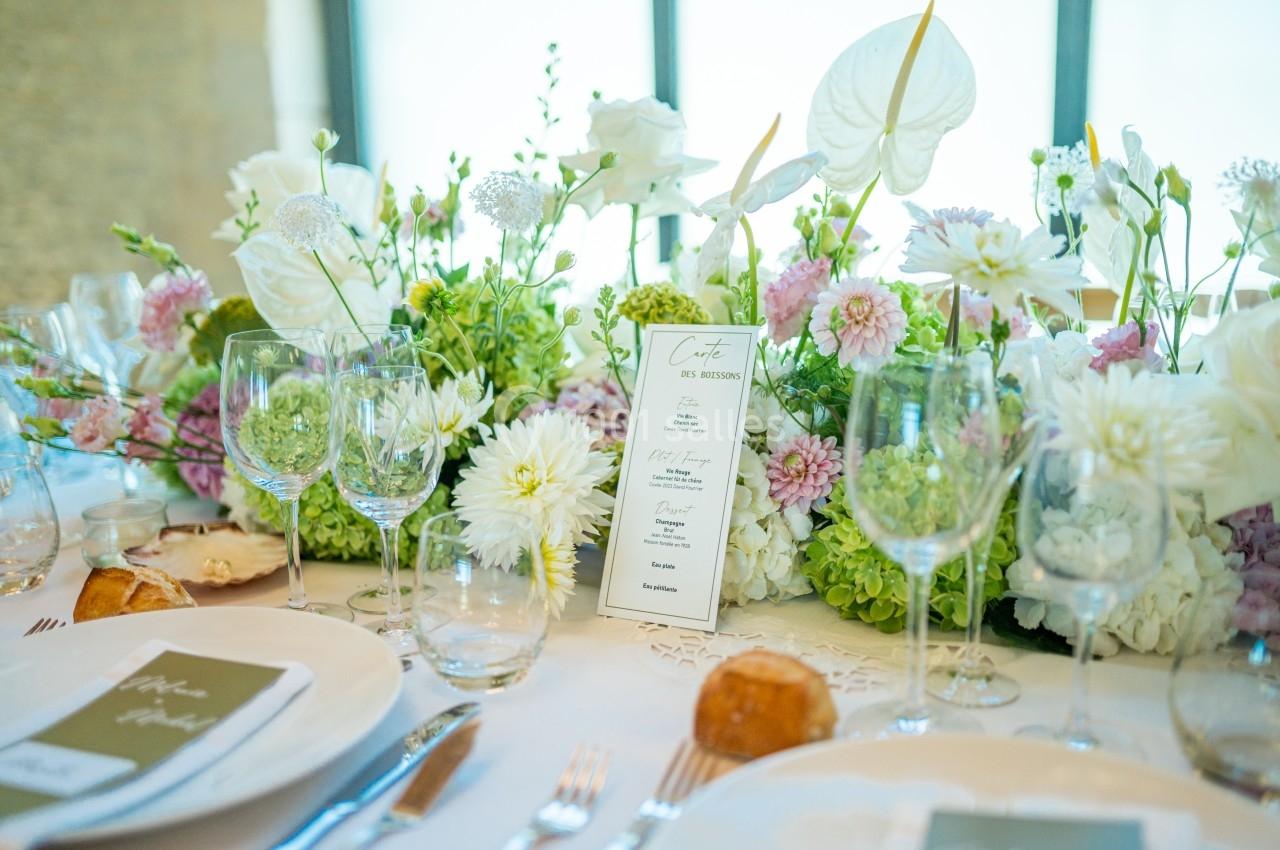 Table élégamment dressée avec des assiettes, verres, menus et un centre de table floral aux tons blancs et verts.