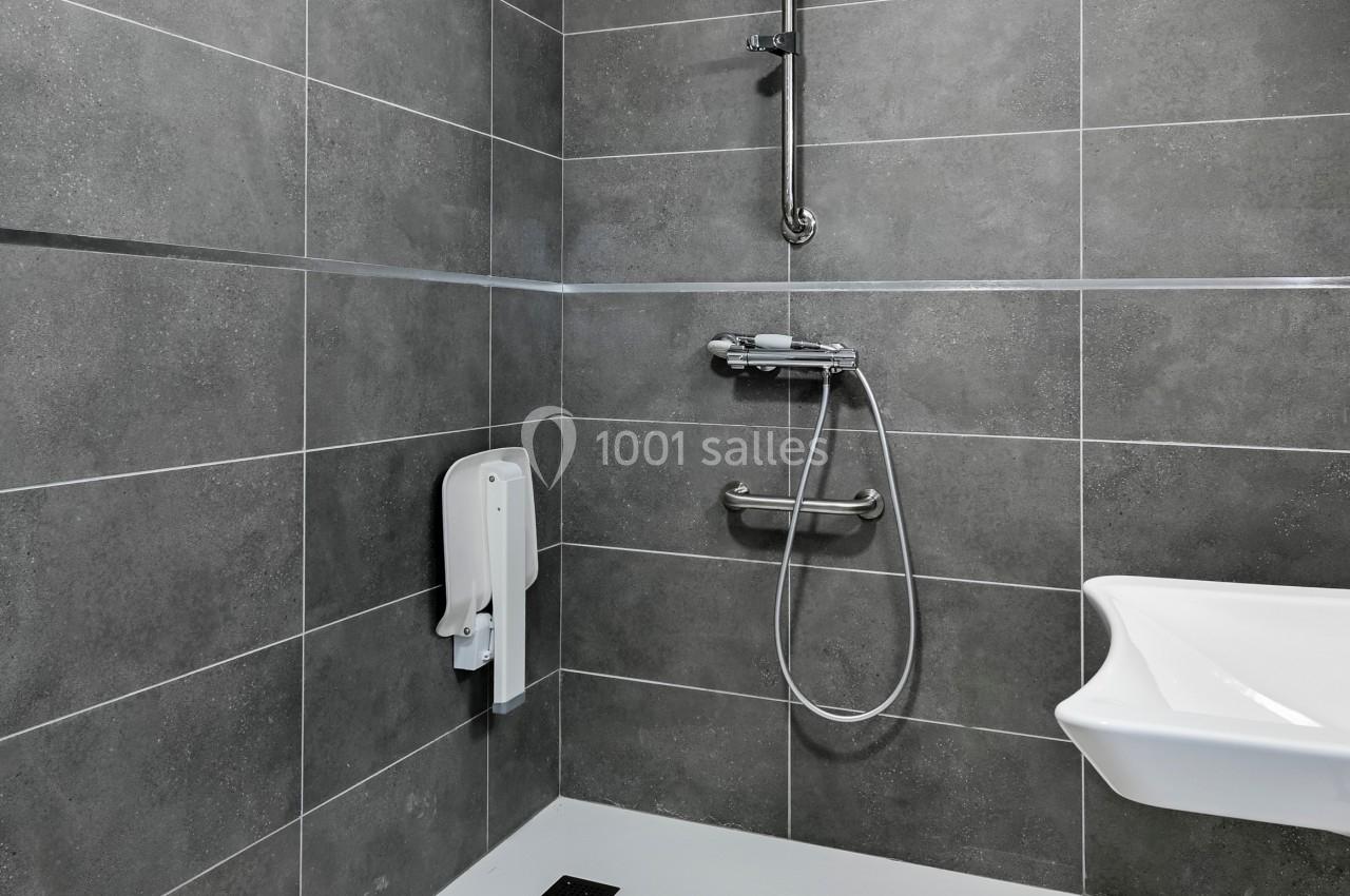 Douche à l'italienne avec carrelage gris, barre d'appui, pommeau réglable et distributeur mural dans une salle de bain.
