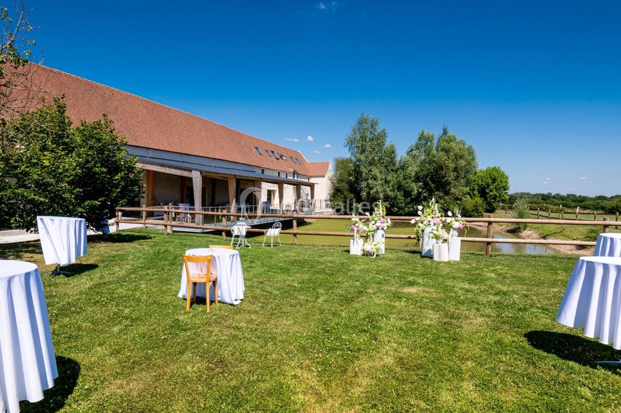 Jardin avec tables hautes dressées, bordé d'un bâtiment en pierre et d'arbres sous un ciel bleu.