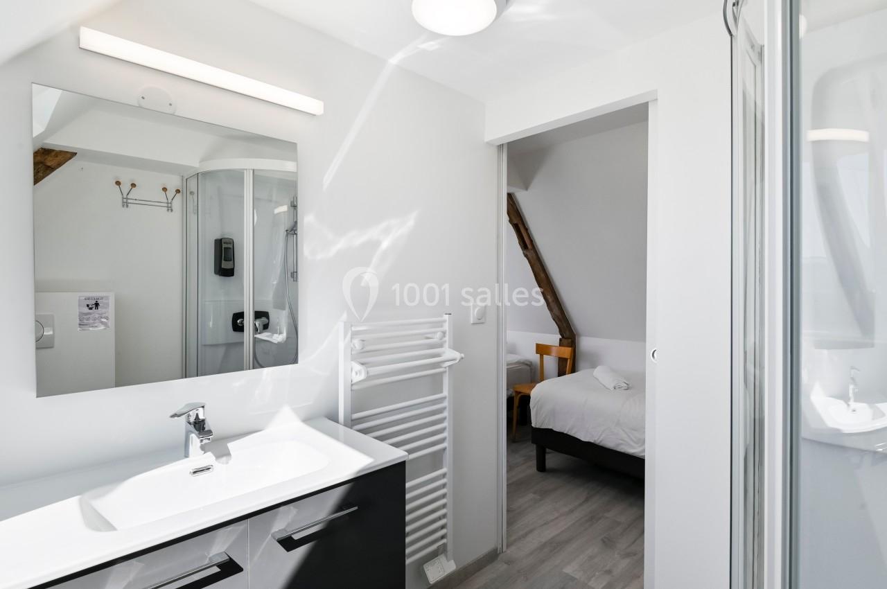 Salle de bain lumineuse avec lavabo, miroir, sèche-serviettes et vue sur une chambre avec lit à travers une porte ouverte.