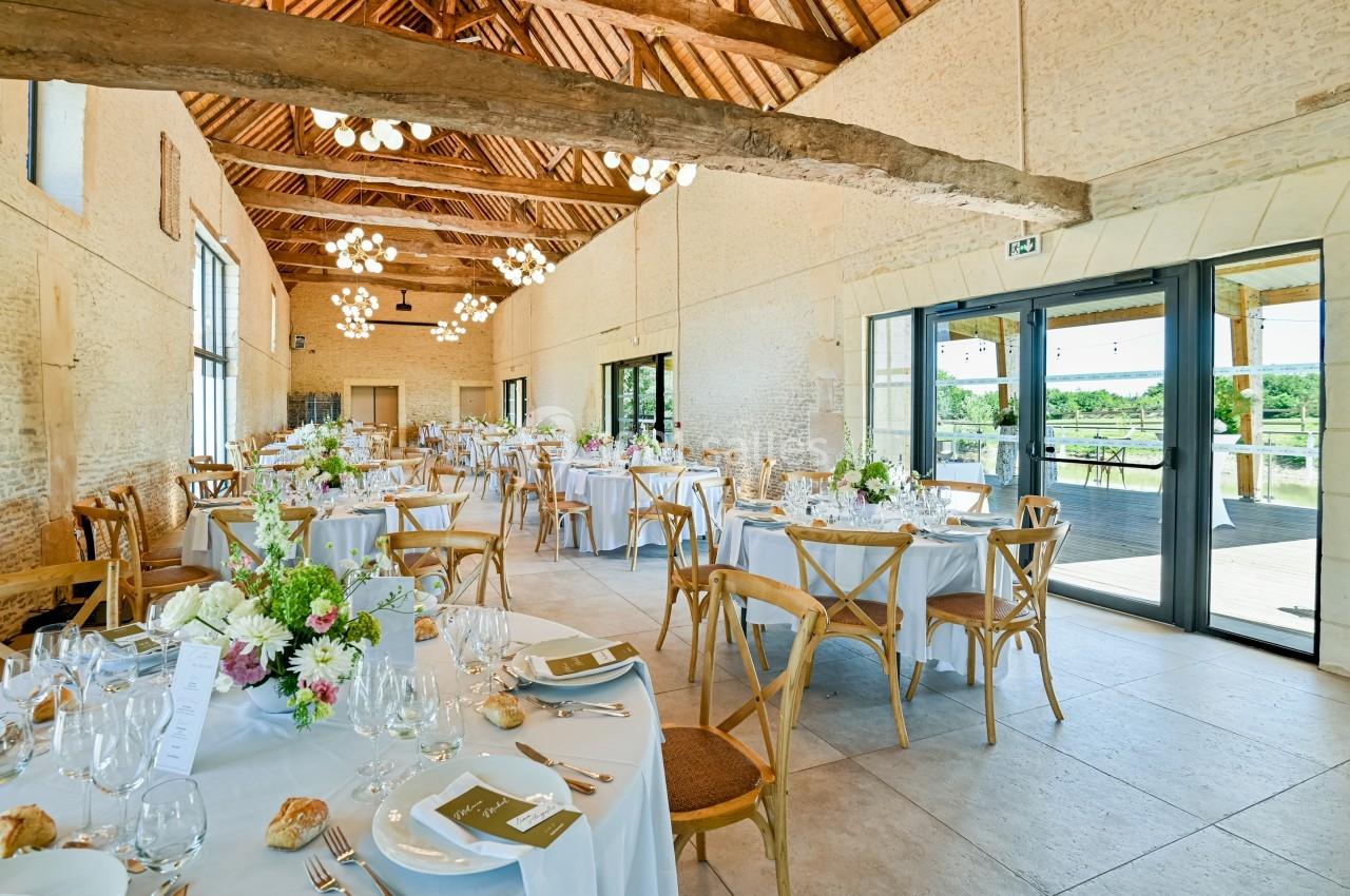 Salle de réception lumineuse avec tables dressées, décorées de fleurs, et vue sur l'extérieur par de grandes baies vitrées.